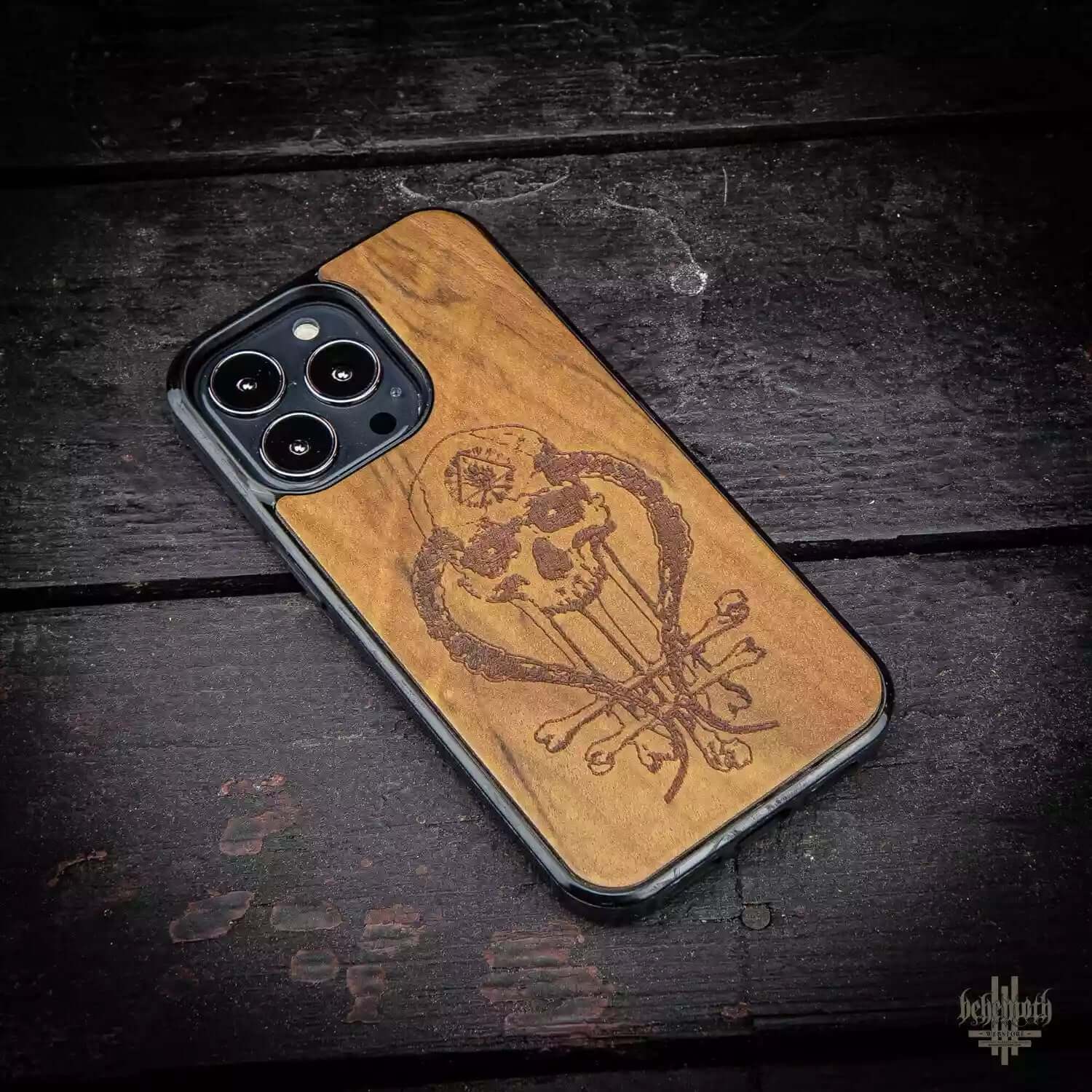 iPhone 13 Pro case with Behemoth 'In Absentia Dei' logo, crafted from exotic Imbuia wood, on a dark wooden table.