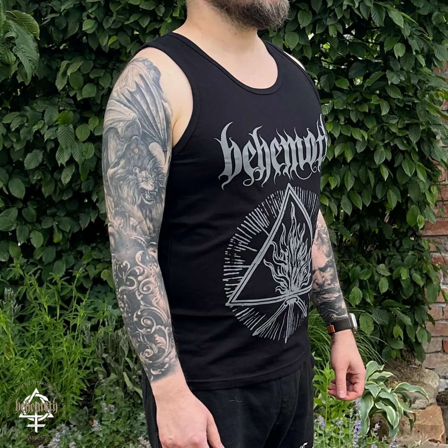 Man wearing black Behemoth 'The Unholy Trinity' tank-top with 'The Satanist' design, standing outside near greenery.