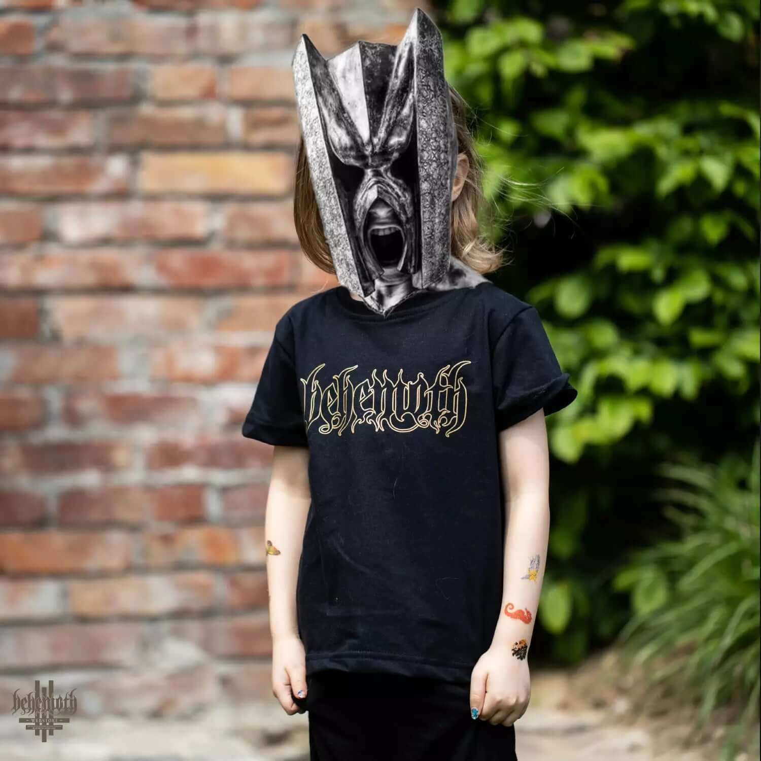 Child wearing a black Behemoth logo t-shirt with safe, OEKO-TEX 100 certified cotton, outdoor scene with brick and greenery background.