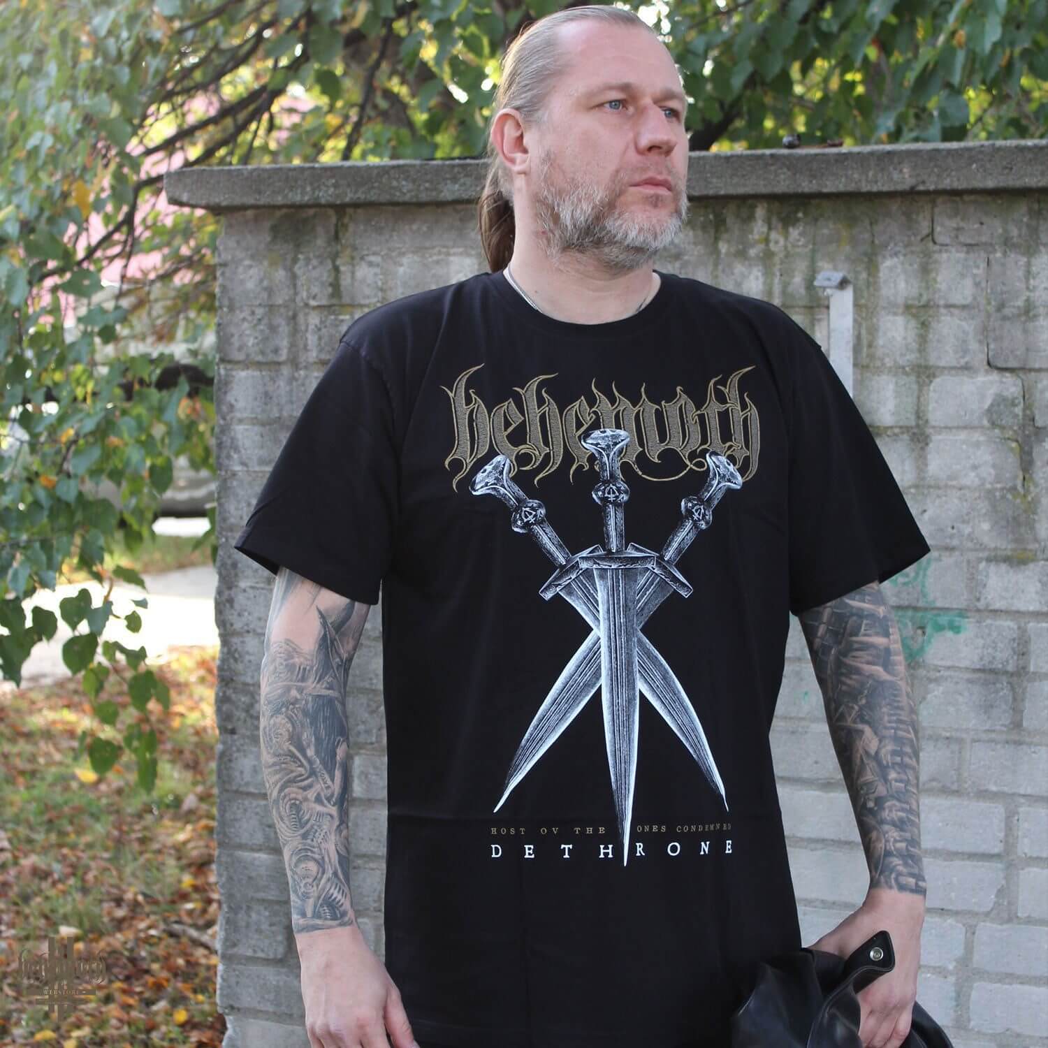 Man wearing 'Off To War' Behemoth T-Shirt with bold graphic design, standing outdoors.