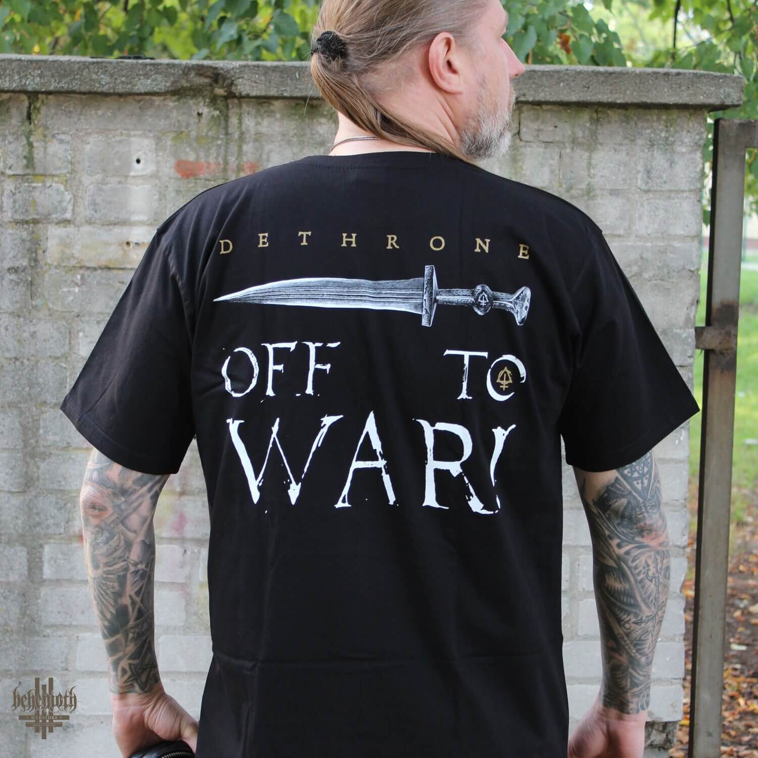 Man wearing 'Off To War' Behemoth T-shirt showcasing back print with sword design and bold lettering in a natural outdoor setting.