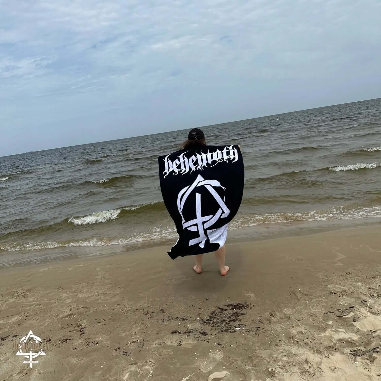 Person holding a Behemoth 'Contra' 100% cotton towel on a beach, showcasing its design and premium quality.