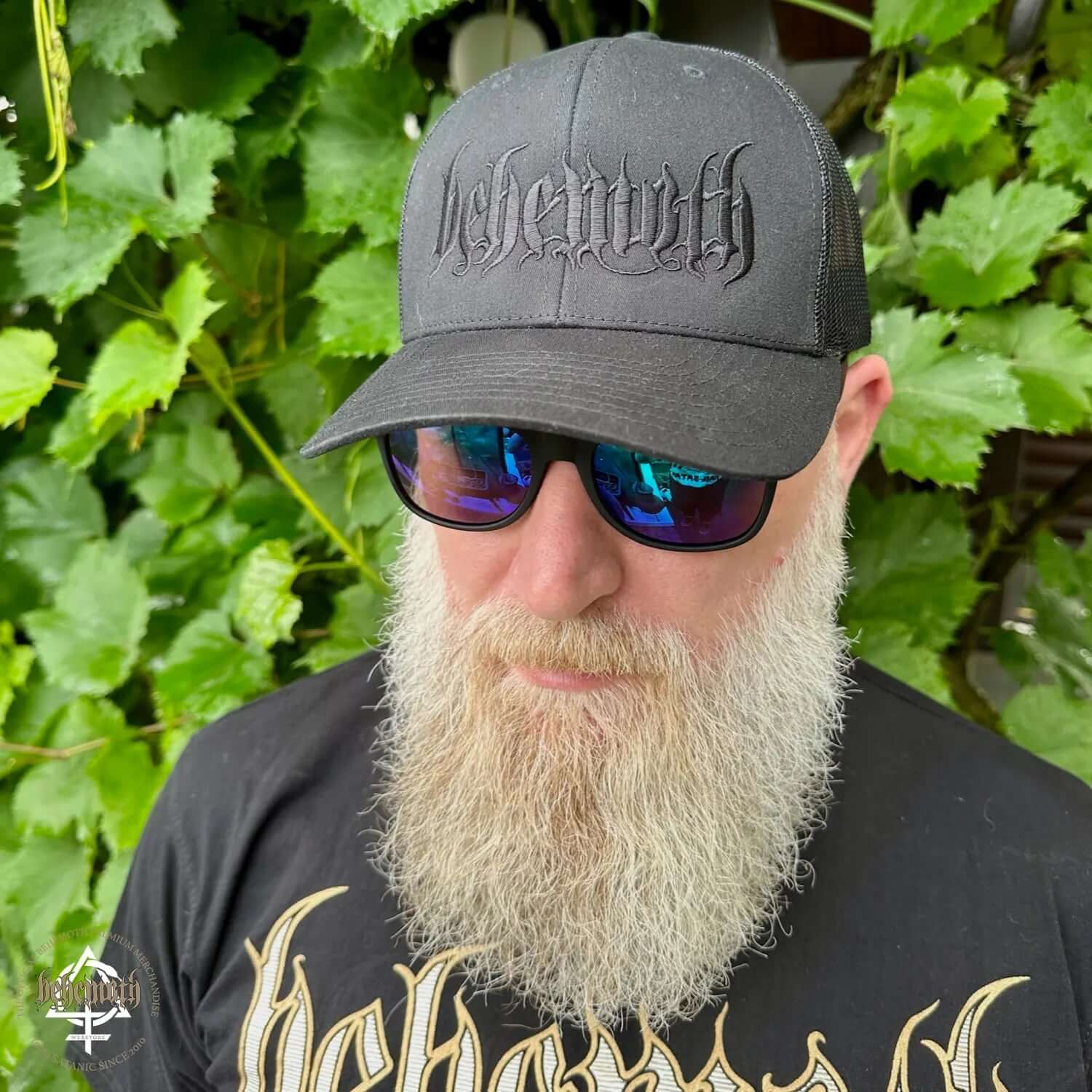 Man wearing Behemoth 'Logo' classic trucker cap BOTB edition with black embroidery, sunglasses, and a matching shirt, standing outside.