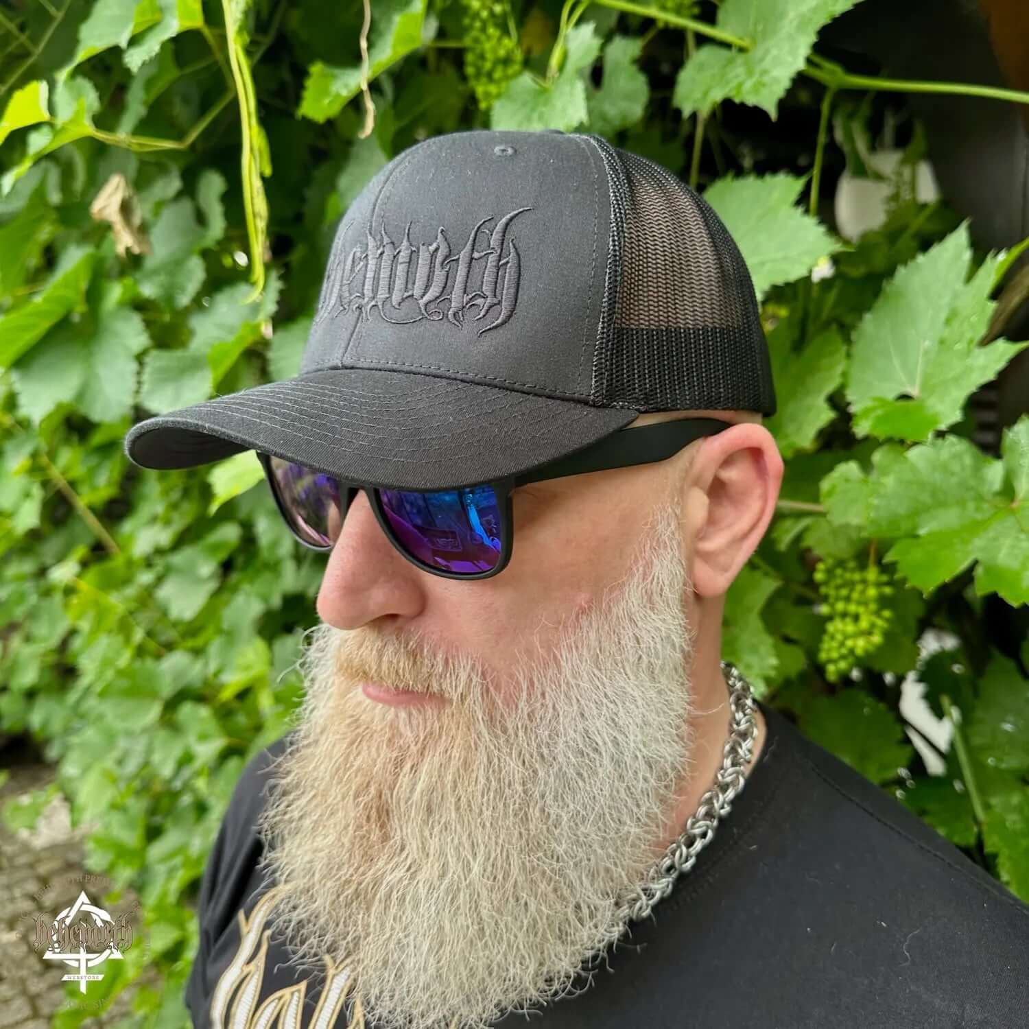 Man wearing a black Behemoth logo trucker cap with sunglasses, standing in front of green foliage.