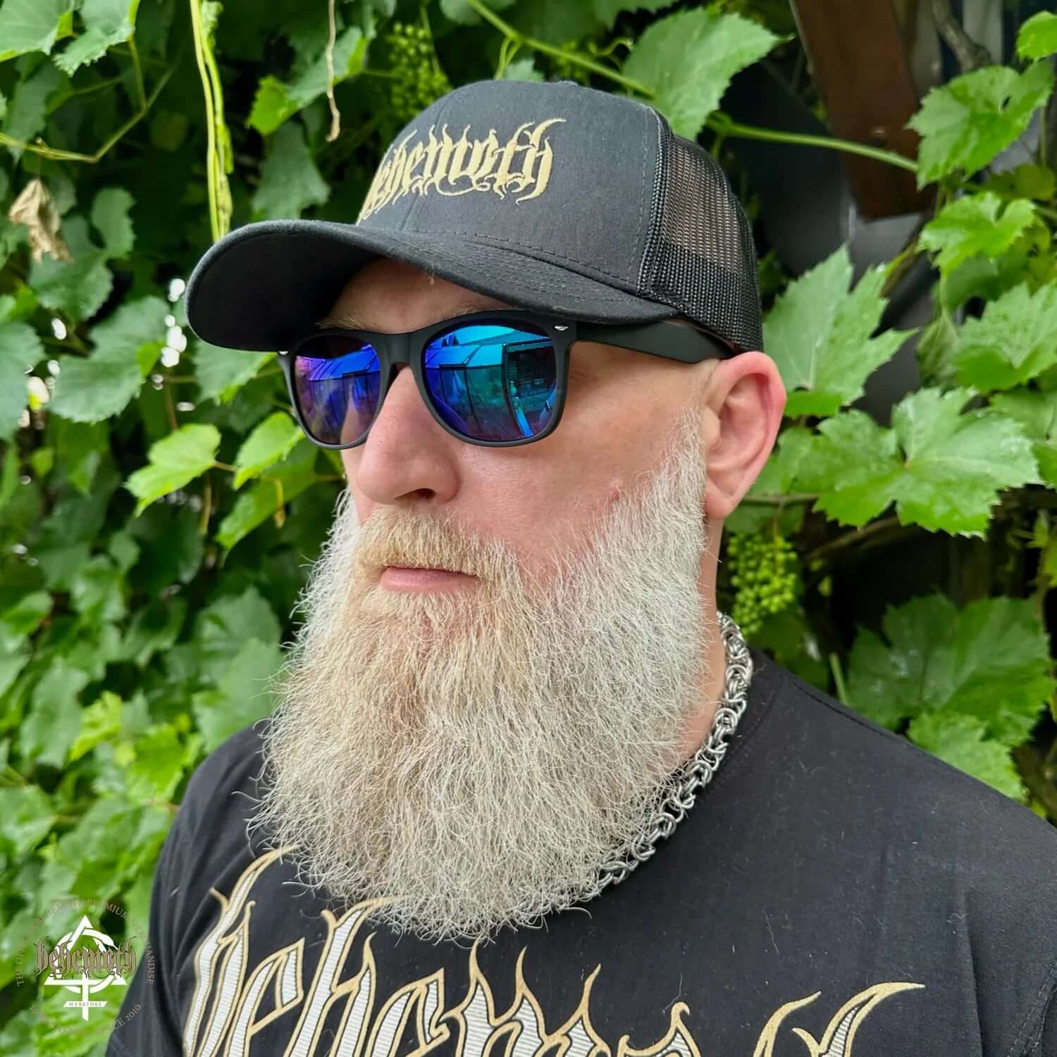 Man wearing Behemoth classic trucker cap with embroidered logo, sunglasses, and chain necklace, standing against green foliage background.
