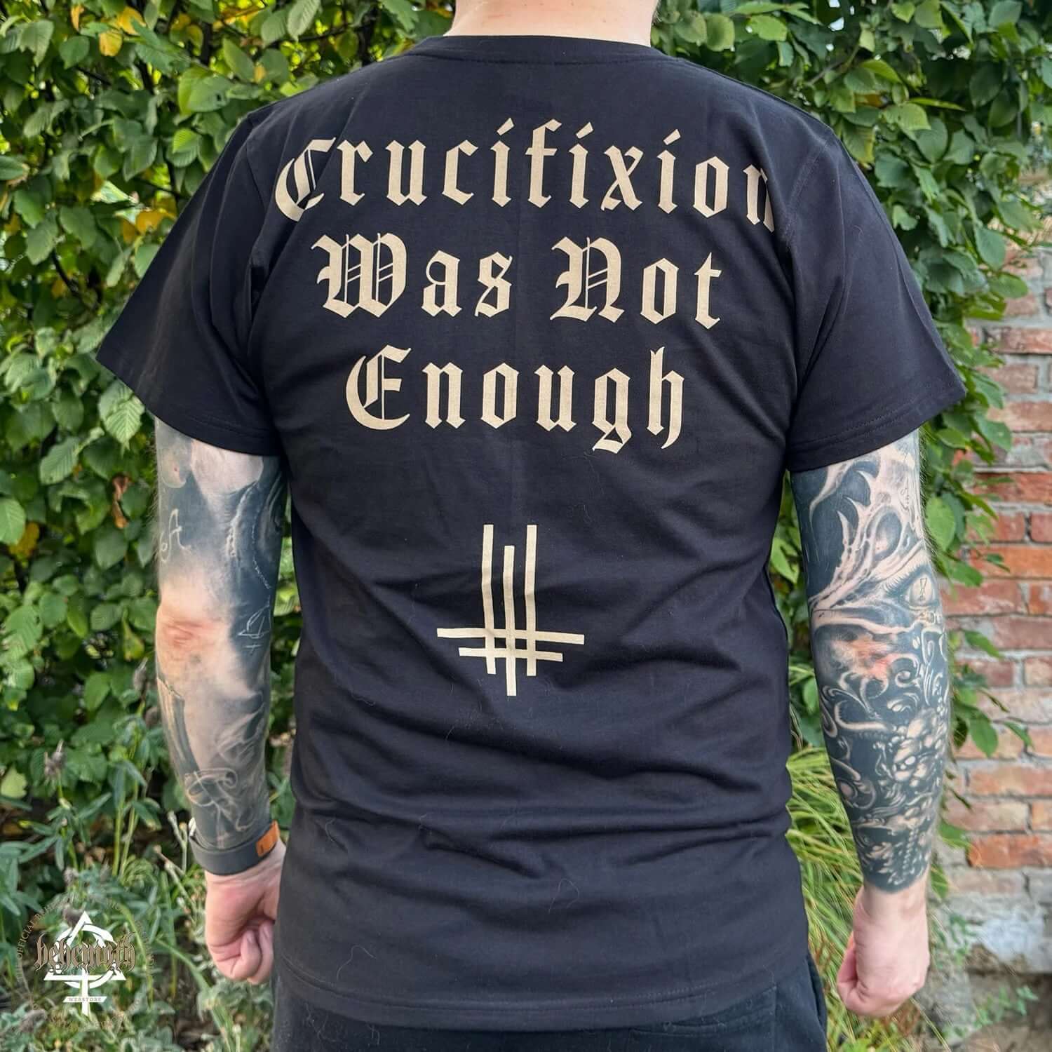 Back view of 'Crucifixion Was Not Enough' Behemoth T-shirt, black with bold gothic text and symbol, worn outdoors, unisex sizing.