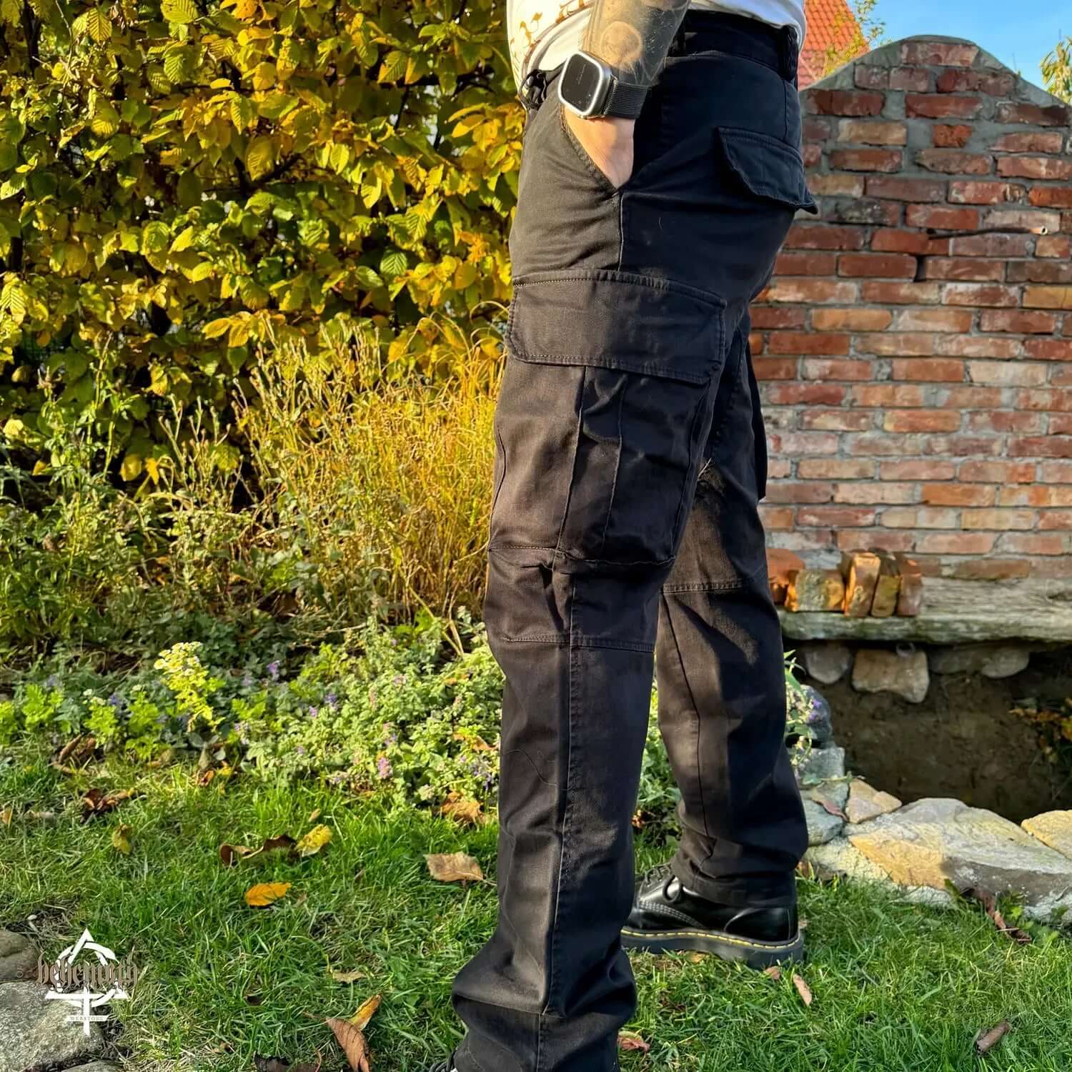 Black Behemoth 'Thru Fire We Walk' cargo trousers with logos, side view, outdoors, showcasing durable material and cargo pockets.