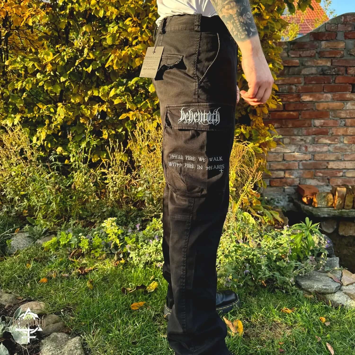 Man wearing Behemoth 'Thru Fire We Walk' black cargo trousers with embroidered logos, standing in a garden.