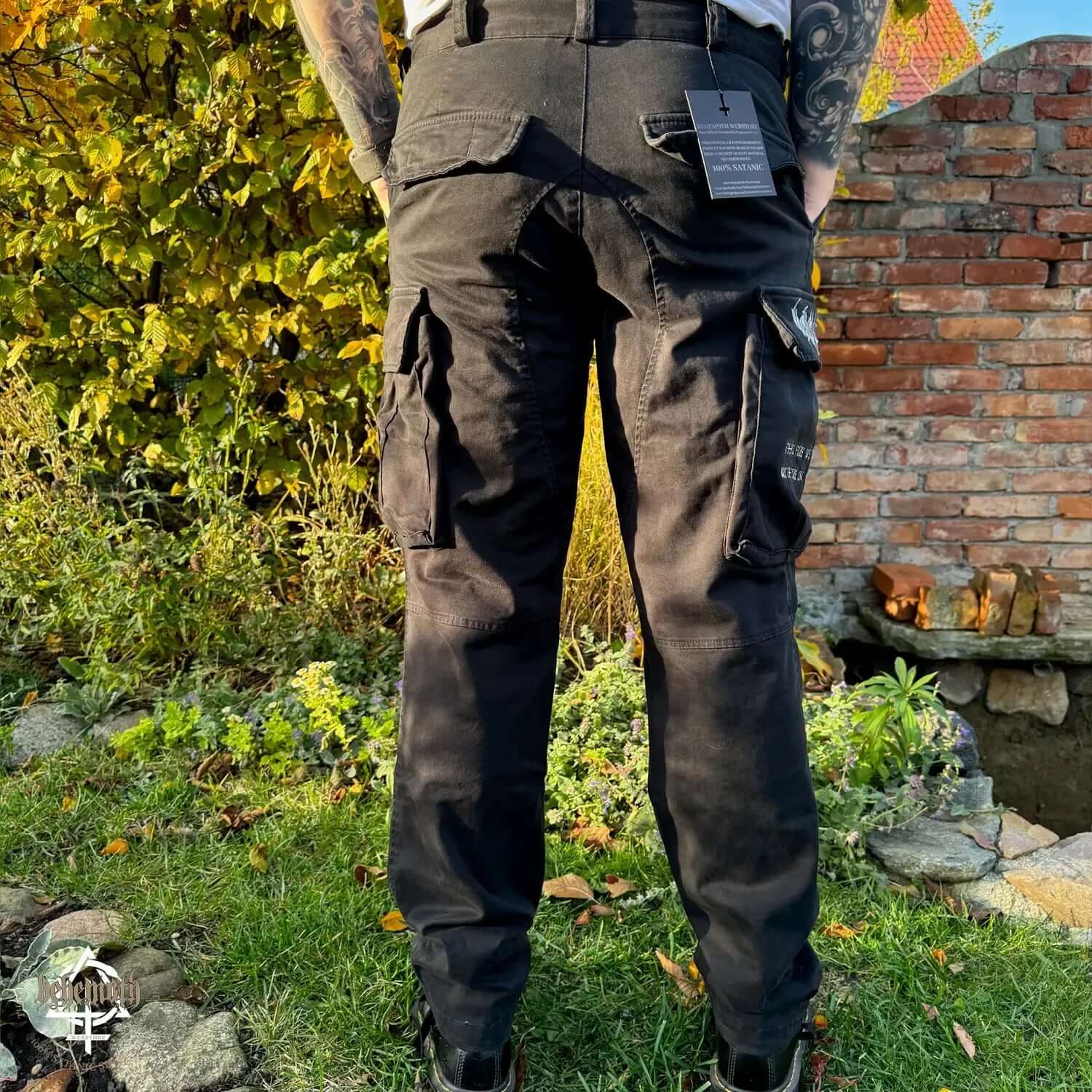 Back view of Behemoth 'Thru Fire We Walk' black cargo trousers with embroidered logos and thigh pockets, outdoors.
