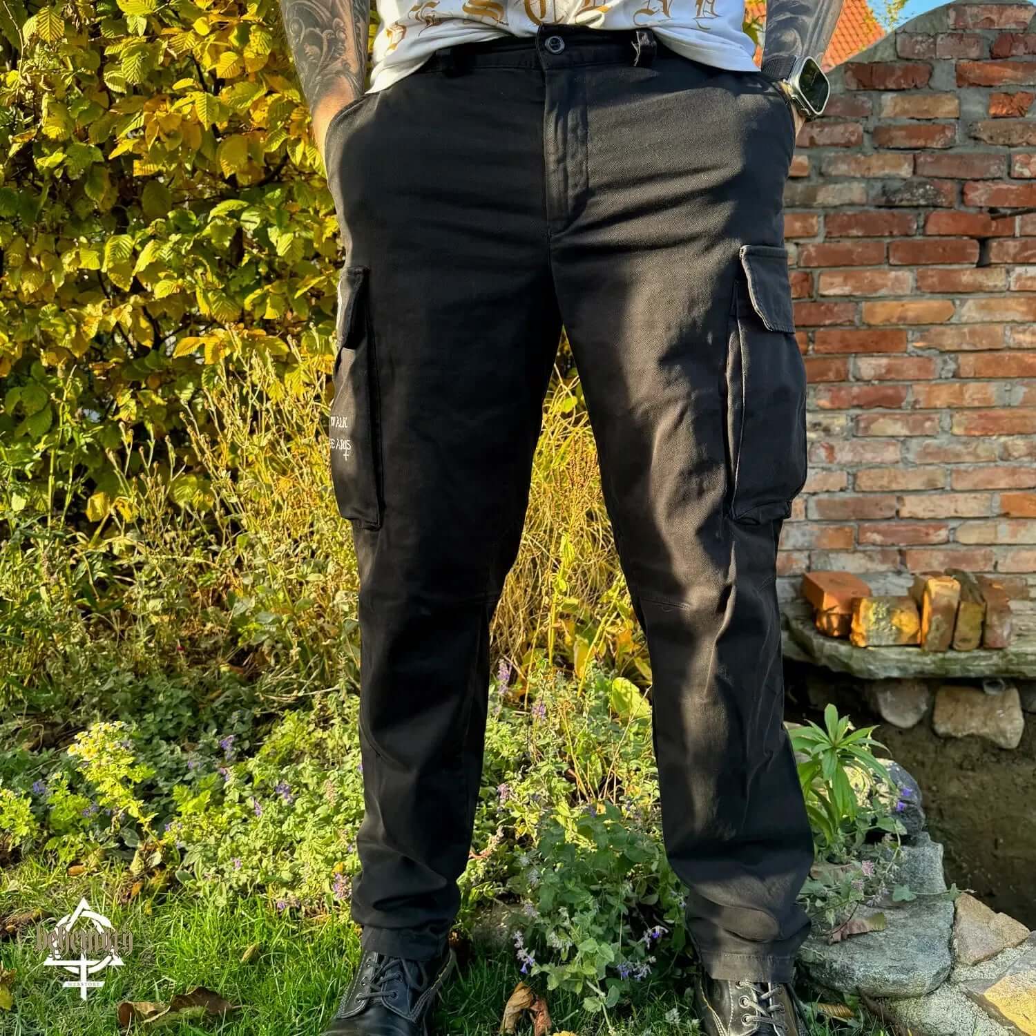 Black 'Thru Fire We Walk' cargo trousers with Behemoth logos, featuring high-quality Polish craftsmanship in a garden setting.