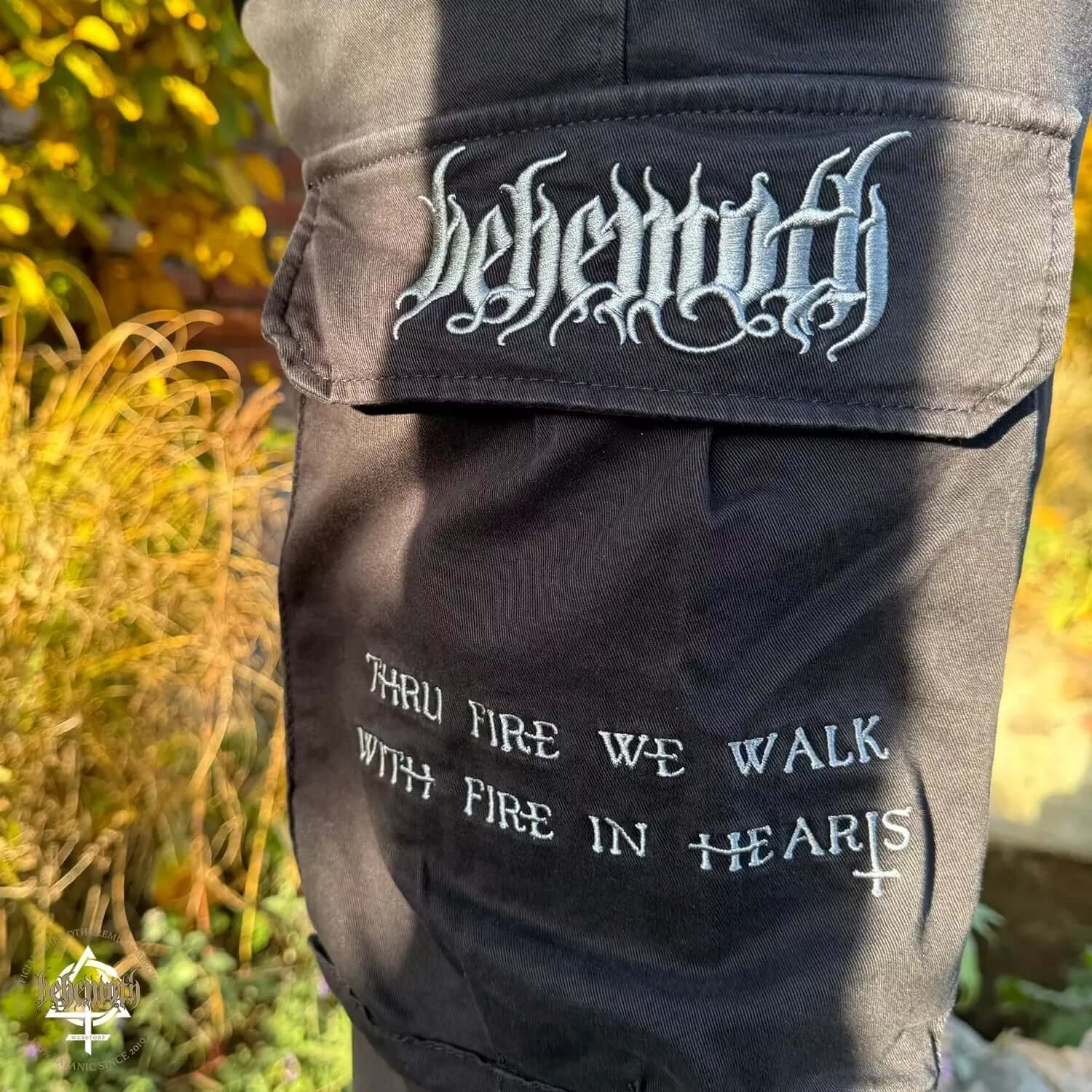 Close-up of Behemoth 'Thru Fire We Walk' black cargo trousers with embroidered logos outdoors
