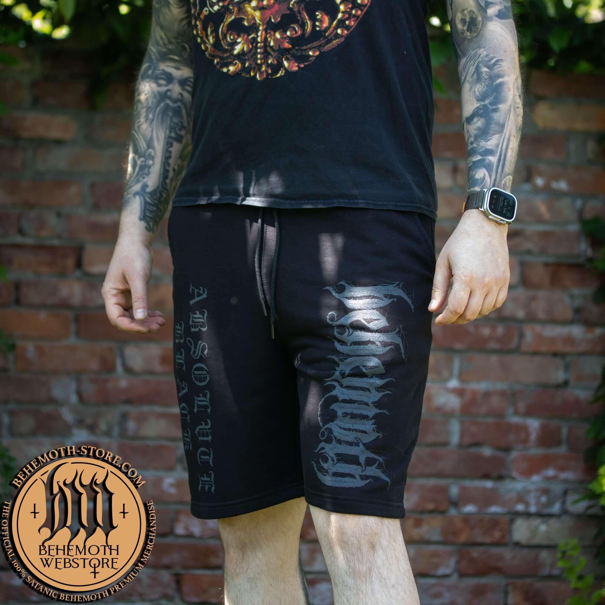Man wearing Behemoth Absolvte Black short sweatpants with screen-printed design, outdoor setting.