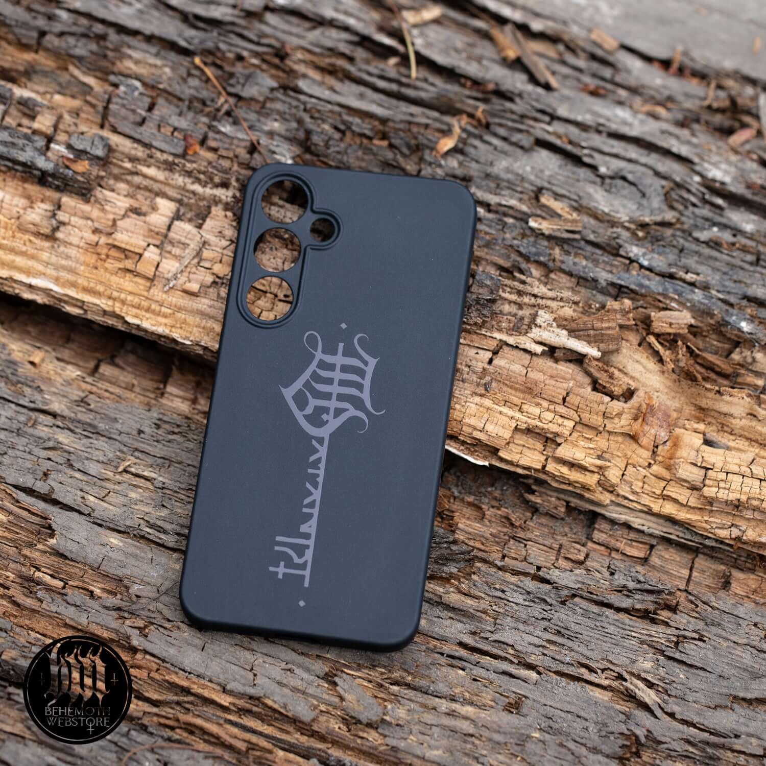 Black matte Behemoth 'The Satanist' phone case with glossy key sigil, designed for optimal grip and phone protection on a wooden surface.