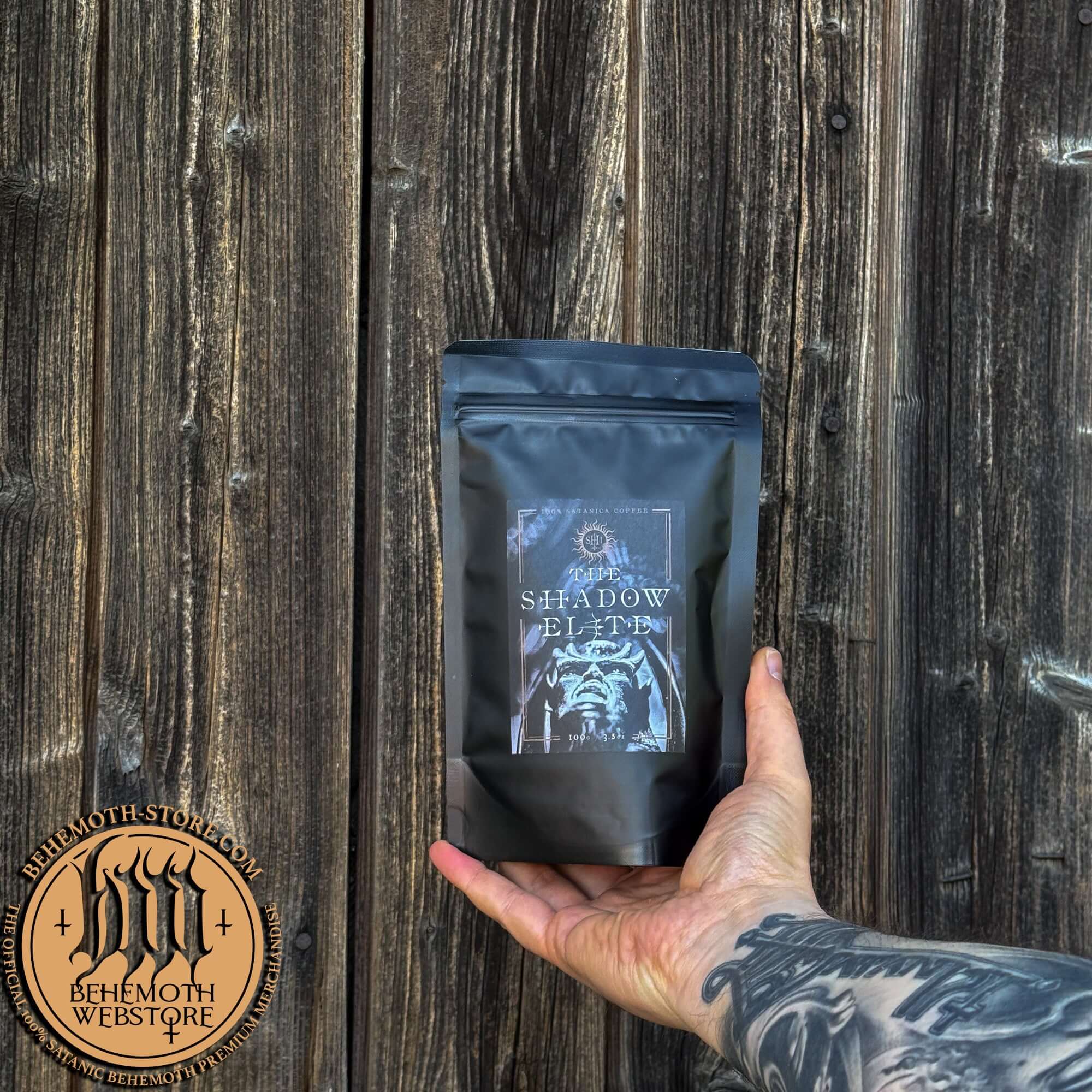 Behemoth 'The Shadow Elite' specialty coffee whole beans package held in tattooed hand against wooden background.