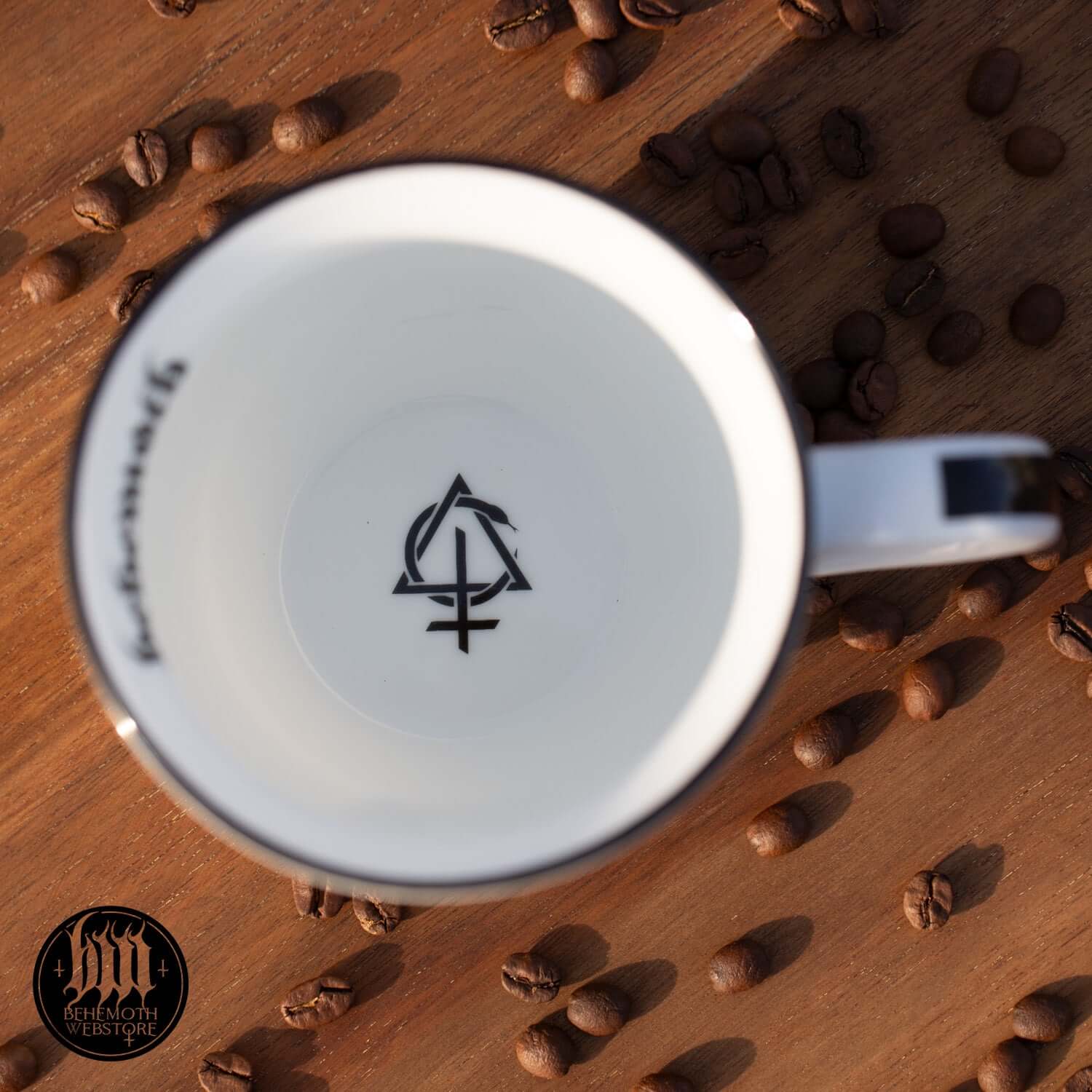 Overhead view of Behemoth 'Polonaise Satanique' mug with symbol inside and coffee beans scattered on wooden surface.