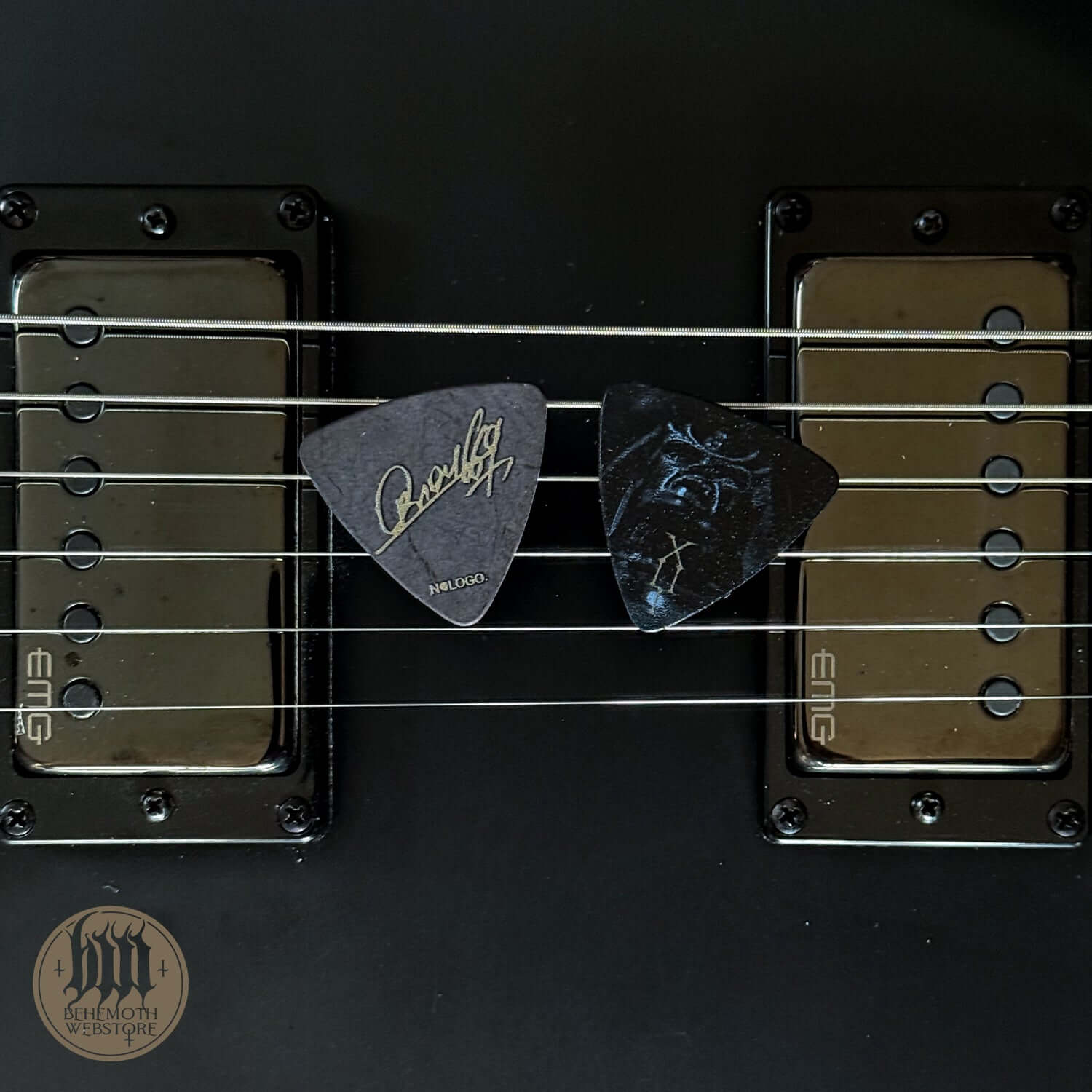Two guitar picks, including Orion signature 'TSOG 13', resting on electric guitar strings over dual EMG pickups.